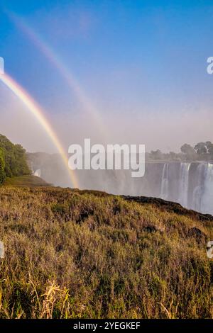 Un vibrante doppio arcobaleno si innalza sullo spruzzo nebbioso delle Cascate Vittoria, creando uno spettacolo naturale mozzafiato all'alba, durante la stagione secca. Foto Stock