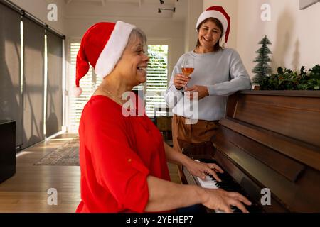 Natale, amiche anziane che indossano cappelli di Babbo Natale, ascoltano musica al pianoforte e vino a casa Foto Stock