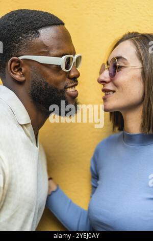 Foto in primo piano verticale di una coppia multietnica moderna e felice con occhiali da sole che stanno per baciarsi su sfondo giallo Foto Stock