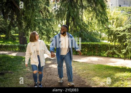 Felice coppia multietnica che indossa abiti casual in denim che camminano tenendosi per mano in un parco Foto Stock