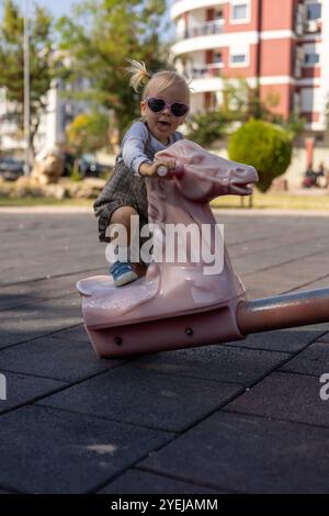 I bambini piccoli amano cavalcare un cavallo a dondolo rosa in un parco giochi soleggiato durante il pomeriggio Foto Stock