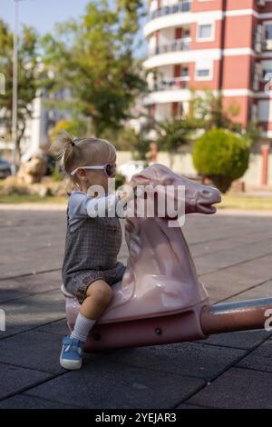 I bambini piccoli amano cavalcare un cavallo a dondolo rosa in un parco giochi soleggiato durante il pomeriggio Foto Stock