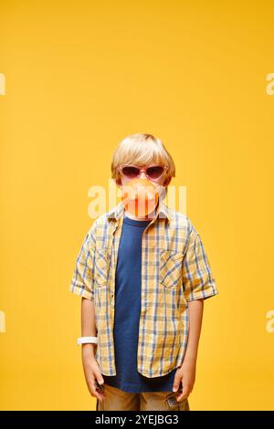 Ripresa verticale di un bambino con occhiali da sole che soffiano bolle d'aria mentre si trova sullo sfondo giallo in studio, copia spazio Foto Stock