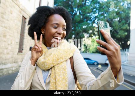 Donna gioiosa che porta Selfie all'aperto con sciarpa gialla e segno di pace in un vivace ambiente urbano Foto Stock