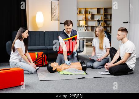 Istruttore di giovane uomo che aiuta a fare le compressioni di cuore di primo soccorso con il dummy durante l'addestramento di gruppo all'interno. Foto Stock