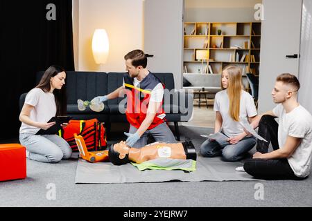 Istruttore di giovane uomo che aiuta a fare le compressioni di cuore di primo soccorso con il dummy durante l'addestramento di gruppo all'interno. Foto Stock