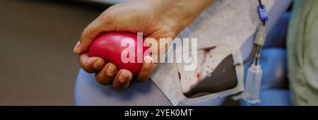 Mano che tiene la palla a forma di cuore mentre donano il sangue tramite gocciolamento endovenoso. Primo piano che mostra la procedura medica e il dispositivo utilizzato per la trasfusione di sangue Foto Stock