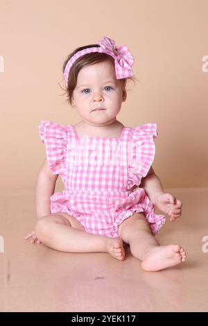 Bellissima bambina dagli occhi azzurri in studio con indosso un abito a scacchi rosa e un archetto abbinato. Adorabile bambina che indossa un abito a scacchi rosa. Foto Stock
