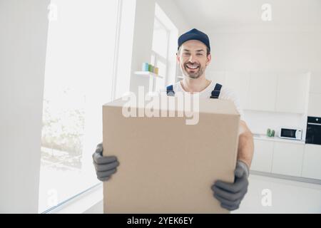 Tutore sorridente che consegna una grande scatola di cartone in un moderno interno della cucina, che mette in mostra un servizio di ristrutturazione professionale con facilità ed efficienza Foto Stock