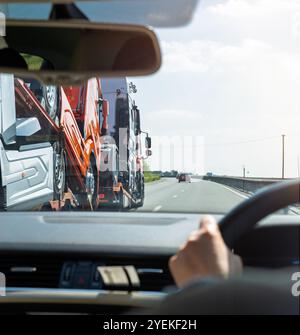 Ripresa dal punto di vista dell'abitacolo di un'auto seguendo un camion che trasporta più camion su un'autostrada, con un cielo limpido e un traffico leggero davanti. Foto Stock