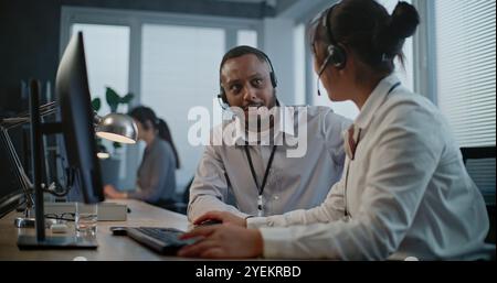 Ufficio del call center: Due operatori multietnici che utilizzano cuffie/auricolari discutono di lavoro e utilizzano il computer per conversare con il cliente su chiamata. Specialisti del supporto tecnico che lavorano nell'assistenza clienti online. Foto Stock