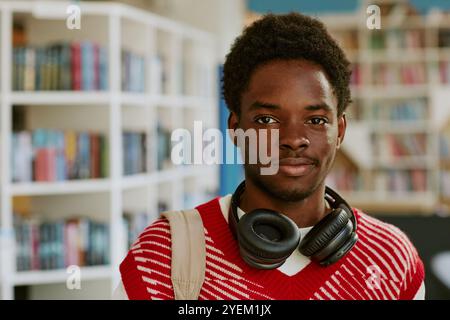 Ritratto di un giovane uomo che indossa un maglione rosso con cuffie intorno al collo in stile biblioteca, che porta con sé uno zaino beige, scaffali pieni di libri sullo sfondo Foto Stock