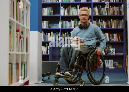 Giovane seduto su una sedia a rotelle che tiene in mano libri mentre indossa le cuffie in un ambiente di biblioteca. È circondato da scaffali pieni di vari titoli e generi Foto Stock