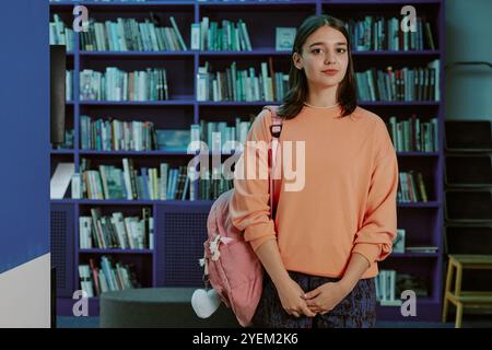 Ritratto di una giovane donna in biblioteca con uno zaino rosa. Scaffali pieni di libri che creano uno sfondo, e lei guarda direttamente alla macchina fotografica Foto Stock