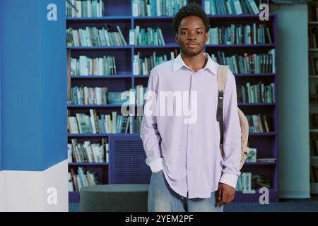 Ritratto di un giovane afroamericano in piedi in una moderna biblioteca con scaffali in sottofondo, vestiti informali e zaino Foto Stock