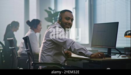Operatore del supporto tecnico afroamericano in cuffia, seduto davanti al computer, guarda la fotocamera che fornisce assistenza clienti. Un team di diversi specialisti dell'helpdesk lavora nel moderno ufficio del call center. Foto Stock