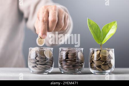 La mano getta le monete in un vaso di vetro, con pile di monete disposte dal più piccolo al più grande sul tavolo. Lo sfondo grigio enfatizza il concetto di Foto Stock