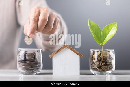 L'uomo getta le monete in un vaso di vetro, con pile di monete e un modello di casa in legno accanto a lui. Enfatizza il concetto di risparmio di denaro per le case future in Foto Stock