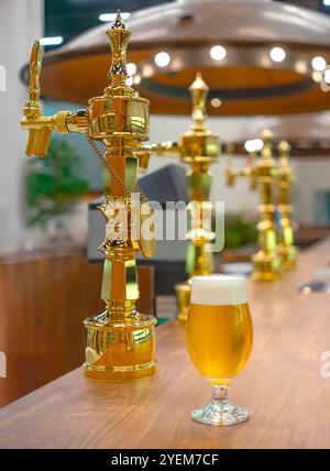 Un bicchiere di birra dorata in tulipano con una testa spumeggiante siede su un bancone bar di legno in una birreria che presenta rubinetti dorati decorati catturati con una bassa profondità Foto Stock