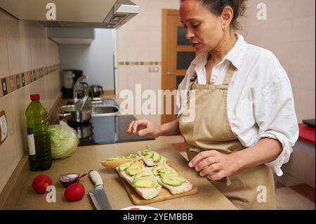 Una donna in grembiule prepara panini all'avocado in una cucina casalinga. Ingredienti freschi come pomodori e lattuga sono sul bancone, incarnando un condimento sano Foto Stock