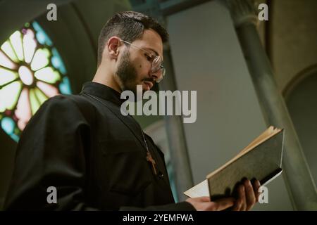 Foto a basso angolo di giovane pastore con bicchieri e vestito in abito con croce di legno sul petto che legge la Bibbia durante la messa in chiesa locale Foto Stock