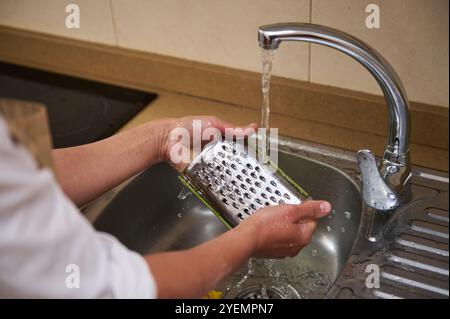 Primo piano delle mani che puliscono una grattugia metallica sotto l'acqua corrente. Il processo di lavaggio degli utensili da cucina dopo l'uso, promuovendo la pulizia e l'igiene Foto Stock