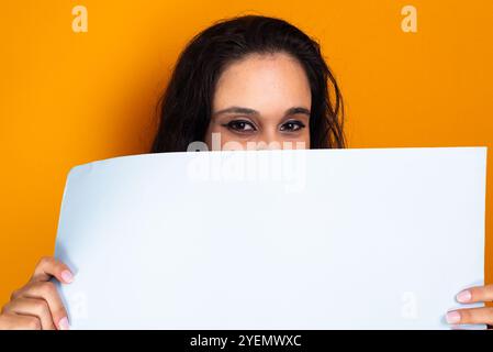 Donna che tiene un poster vuoto davanti al viso. Ripresa in studio con sfondo giallo. Foto Stock