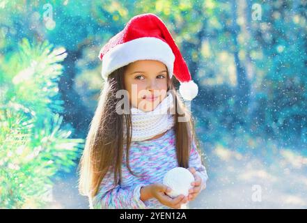 Bambino di Natale bambina in cappello rosso di babbo natale con palla di neve vicino a un albero di ramo Foto Stock