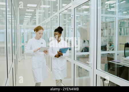Due professionisti del settore medico si impegnano in una discussione mentre camminano attraverso un corridoio con pareti in vetro. Un infermiere sta utilizzando una tavoletta mentre l'altro sta tenendo le cartelle paziente Foto Stock