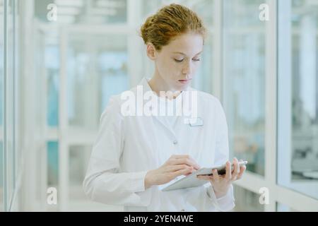 Professionista sanitario femminile in ambiente di laboratorio che esamina i dati su un dispositivo tablet mentre si concentra sull'attività, con background delle apparecchiature mediche e delle impostazioni di laboratorio Foto Stock