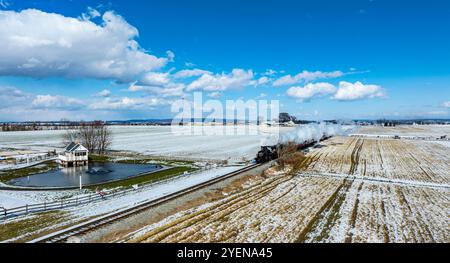 Una locomotiva a vapore si muove lungo i binari, rilasciando vapore mentre si muove attraverso campi innevati. In primo piano, un gazebo si trova vicino a un piccolo po Foto Stock