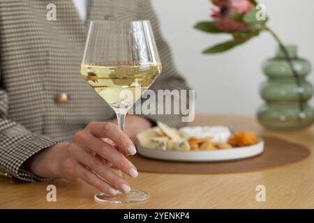 Un bicchiere di vino bianco in mano sullo sfondo di un piatto di formaggi con spuntini e noci Foto Stock