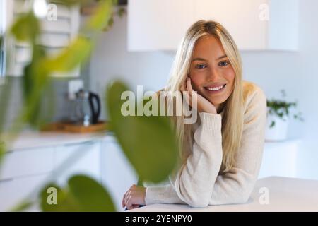 Donna sorridente in un comodo maglione appoggiato sul bancone della cucina di casa. Stile di vita, relax, informale, domestico, tempo libero, allegro Foto Stock