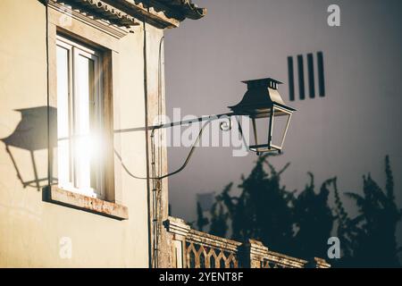 La calda luce del sole si riflette su una applique in stile vintage montata sul lato di un vecchio edificio in stucco con una grande finestra. crea ombre e luce soffusa Foto Stock