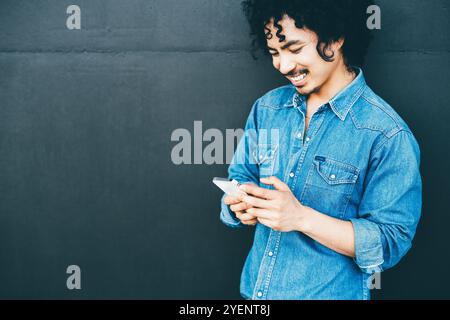 Giovane asiatico in camicia di jeans sorridente mentre utilizza uno smartphone, in piedi su uno sfondo scuro. Sembra concentrato e gioioso; con uno spazio di copia ar Foto Stock