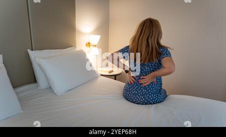 Donna seduta sul letto con le mani dietro la schiena a causa del dolore posturale che ha. Foto Stock