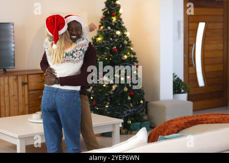 Coppia multirazziale sorridente che si abbraccia all'albero di Natale a casa, indossando cappelli di Babbo Natale. Famiglia, festeggiamenti, festivi, stagionali, amore Foto Stock