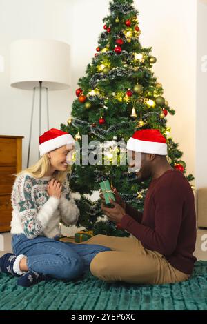 Coppia multirazziale che si scambia regali con l'albero di Natale, indossa cappelli di Babbo Natale, si sente gioiosa, a casa. Festeggiamenti, vacanze, insieme, romanticismo, mestieri Foto Stock