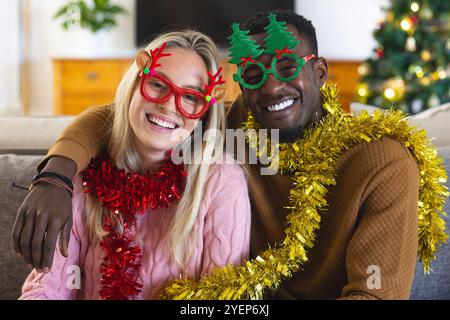 Coppia multirazziale che indossa occhiali e tinsel per festeggiare il Natale a casa. Vacanze, festeggiamenti, felicità, famiglia, diversità, gioioso Foto Stock