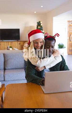 Felice coppia multirazziale che indossa cappelli festivi utilizzando il laptop a casa durante il Natale. Festività, festeggiamenti, digitale, tecnologia, inverno, gioioso Foto Stock