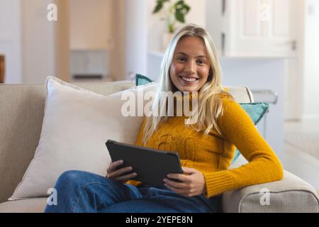 Donna sorridente che si rilassa sul divano con un tablet a casa, godendosi il tempo libero. relax, tecnologia, stile di vita, comfort, digitale, informale Foto Stock
