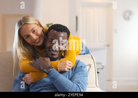 Felice coppia multirazziale che abbraccia l'accogliente divano di casa, godendo le festività natalizie. Celebrazione, felicità, romanticismo, stile di vita, inverno, famiglia Foto Stock