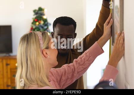 Coppia multirazziale che decora la casa per Natale, appende foto con sorrisi. vacanze, feste, decorazione, insieme, felicità, inverno Foto Stock