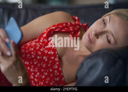 Donna bionda serena a casa, che si rilassa sul divano e utilizza lo smartphone Foto Stock