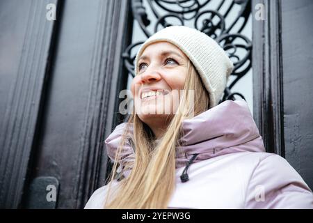 Donna in giacca e cappello invernali sorridente vicino a sfondo ornato all'aperto Foto Stock