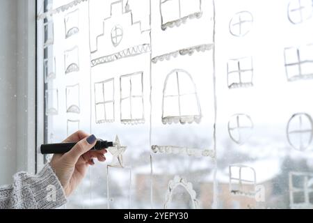 La mano femminile disegna le decorazioni natalizie dipinte su vetro della finestra. Paesaggi di New Years. Edifici della città ghirlande luci. Igge, decorazione e chri Foto Stock