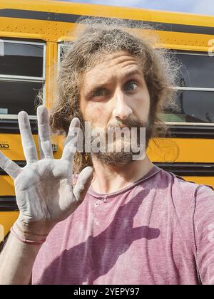 Primo piano ritratto di un giovane dall'aspetto divertente con i capelli lunghi, che mostra una mano sporca spolverata accanto a un autobus in fase di ristrutturazione Foto Stock