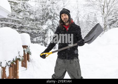 Giovane uomo caucasico con un sorriso, in piedi all'aperto tenendo una pala per spalmare la neve. casa e alberi ricoperti di neve sullo sfondo Foto Stock