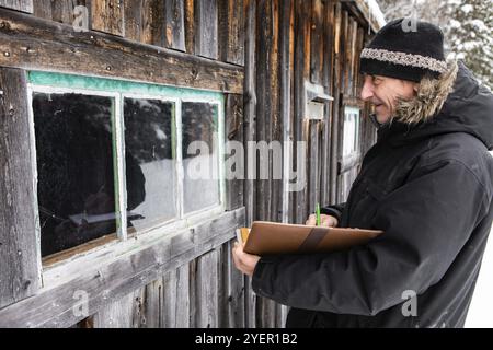 Ispettore domestico di mezza età che prende appunti su un notebook con copertina in pelle durante un'ispezione all'aperto. esaminando una finestra di una vecchia casa di legno Foto Stock
