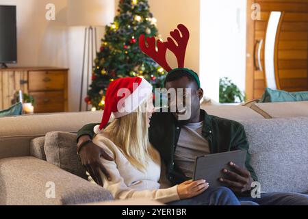 Coppia multirazziale sul divano che festeggia il Natale con un tablet, sorridendo sull'albero. festeggiamenti, tecnologia, festività, famiglia, digitale Foto Stock